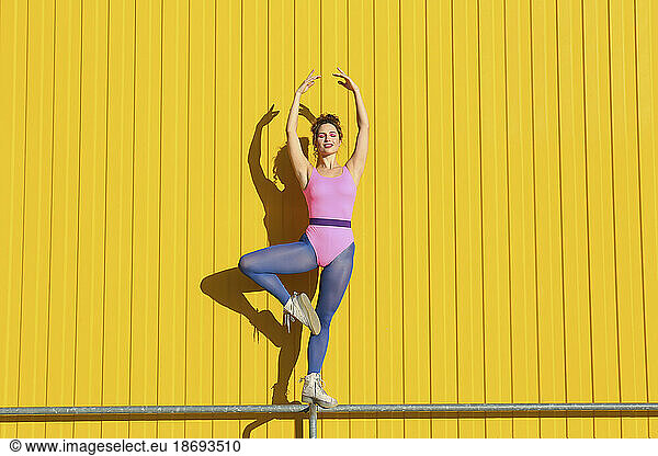 Young woman wearing bodysuit and practicing ballet on railing in front of yellow wall