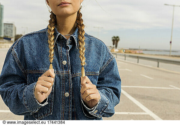 Young woman wearing denim jacket holding braided hair