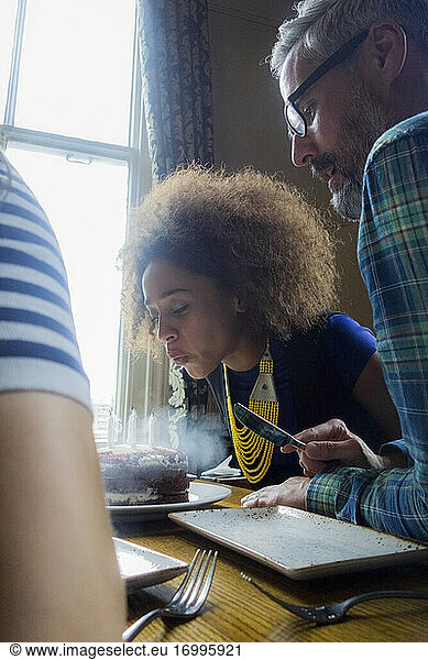 Young woman wearing ethnic jewelry blowing candles on cake while sitting with friends at cafe