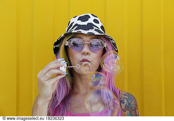 Young woman wearing hat blowing soap bubbles in front of yellow wall