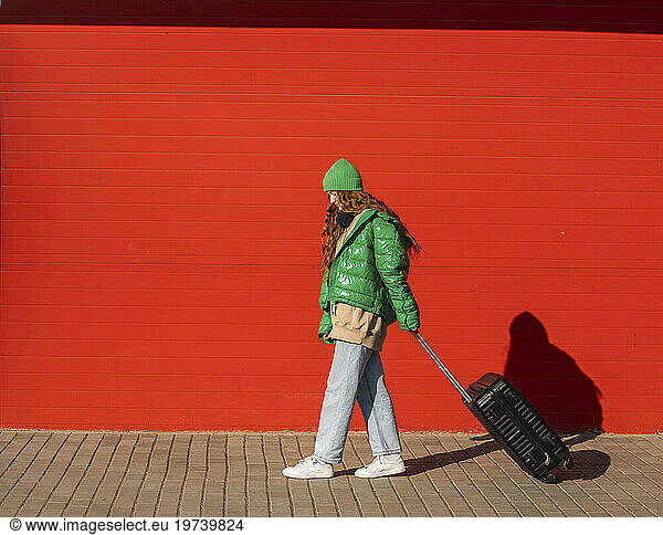 Young woman wearing jacket walking with suitcase in front of red wall