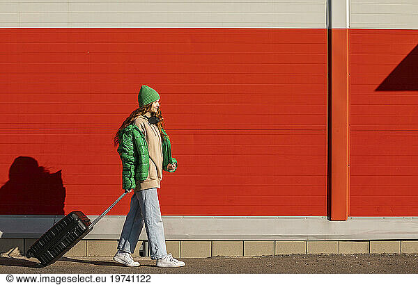 Young woman wearing knit hat walking with wheeled suitcase in front of red wall