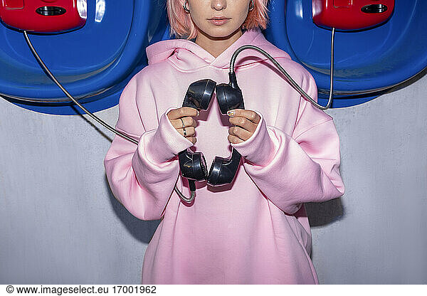 Young woman wearing pink hooded shirt standing in front of telephone booths  holding receivers