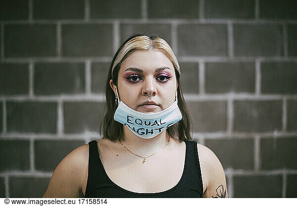 Young woman wearing protective face mask with equal rights text on it against wall