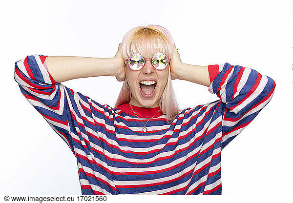Young woman wearing sunglasses covering ears with hands while shouting against white background