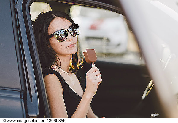 Young woman wearing sunglasses holding frozen sweet food while sitting in car seen through window