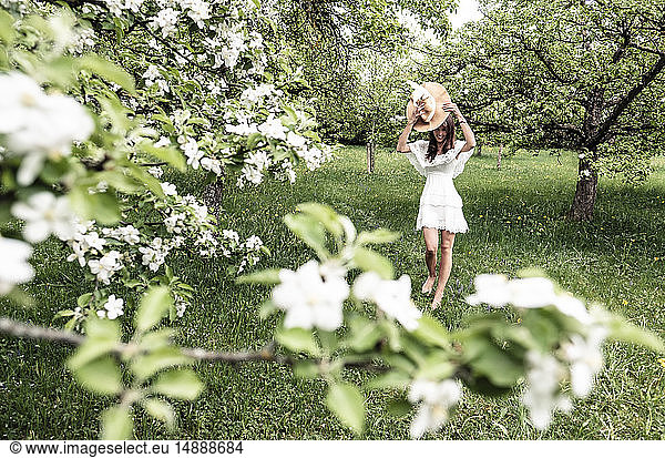 Young woman wearing white dress and floppy hat walking barefoot in garden with blossoming apple trees