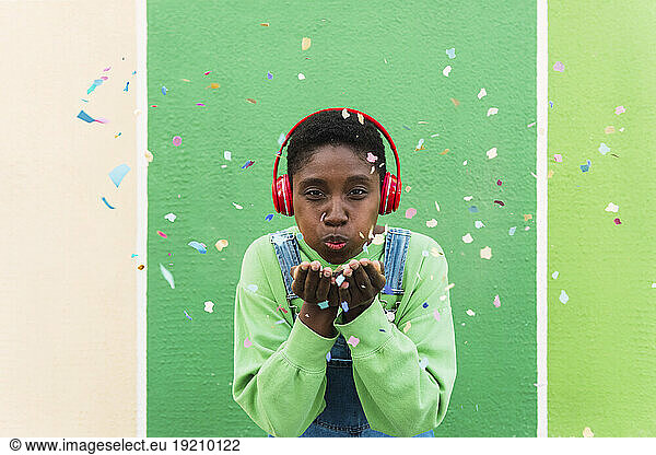 Young woman wearing wireless headphones blowing confetti in front of multi colored wall