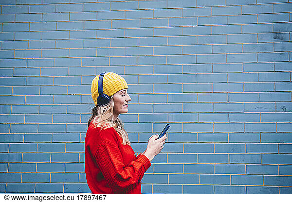 Young woman wearing wireless headphones using smart phone in front of wall