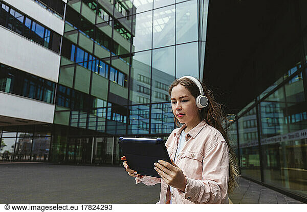 Young woman wearing wireless headphones using tablet PC in front of glass building