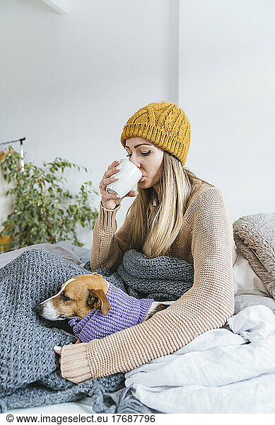 Young woman wearing wooly hat drinkig hot tea sitting in bed with dog