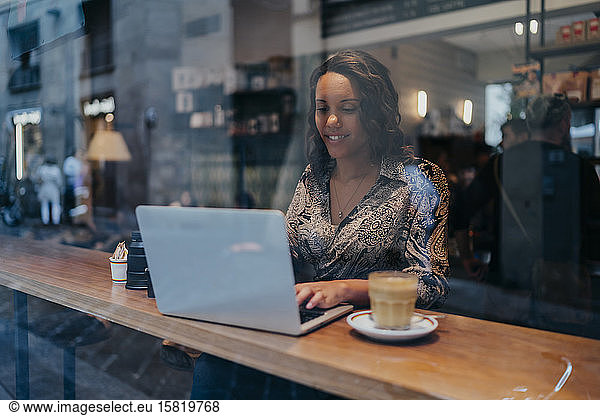 Young woman with a camera using laptop in a cafe behind windowpane