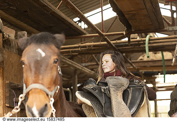young woman with a horse in a horse stable with a saddle