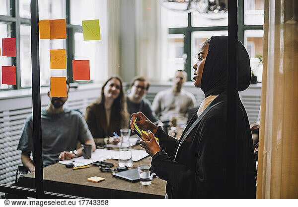Young woman with adhesive notes discussing plan with colleagues during meeting in creative office