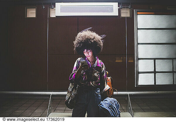 Young woman with afro hairdo using smartphone at bus stop in the city