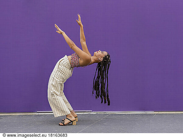 Young woman with arms raised bending over backwards in front of purple wall