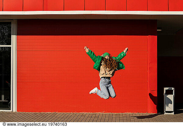 Young woman with arms raised jumping in front of red wall