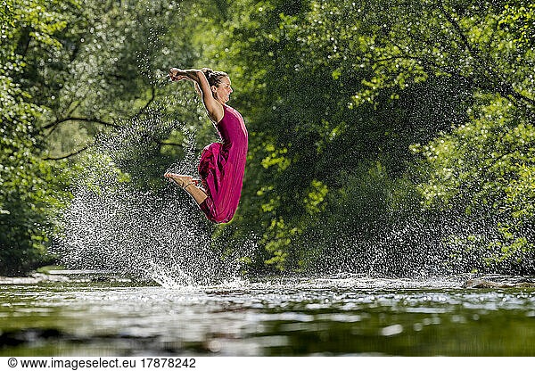 Young woman with arms raised jumping over river