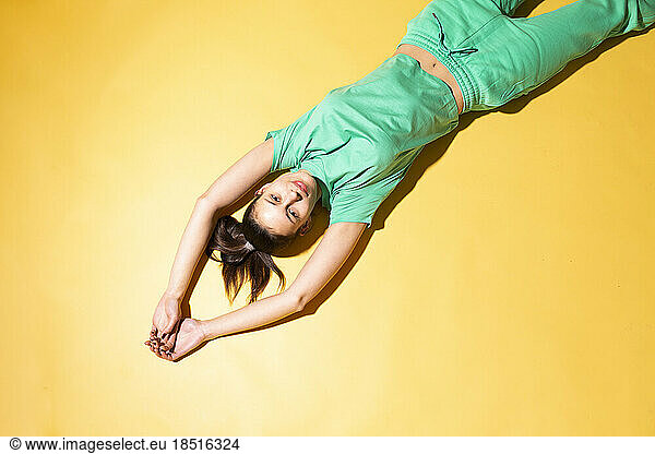 Young woman with arms raised lying on yellow background