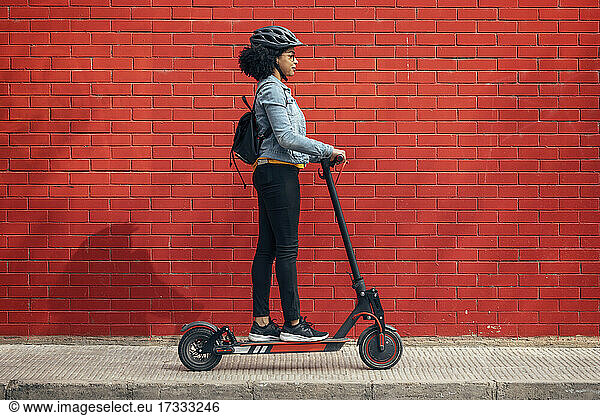 Young woman with backpack riding electric push scooter by red wall