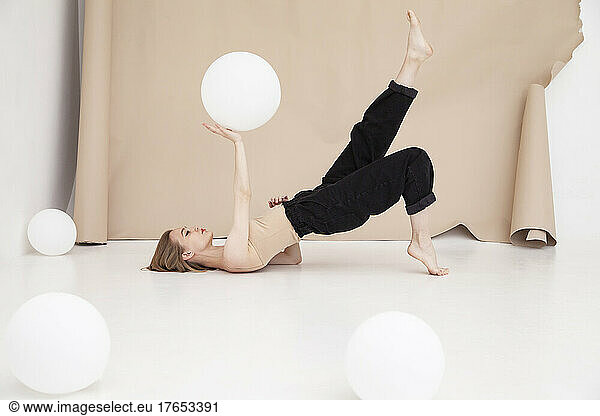 Young woman with balloon on hand exercising yoga by brown backdrop