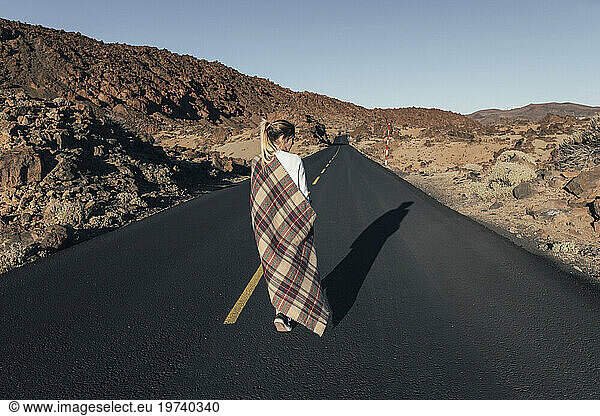 Young woman with blanket walking on road near volcanic landscape
