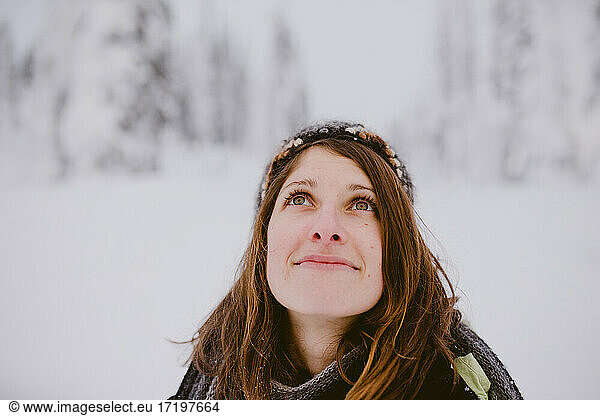 Young woman with brown hair and eyes looks up at snow covered trees