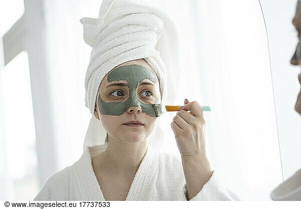Young woman with brush applying facial mask on face at home