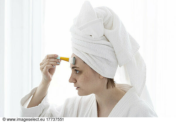 Young woman with brush applying facial mask on forehead at home