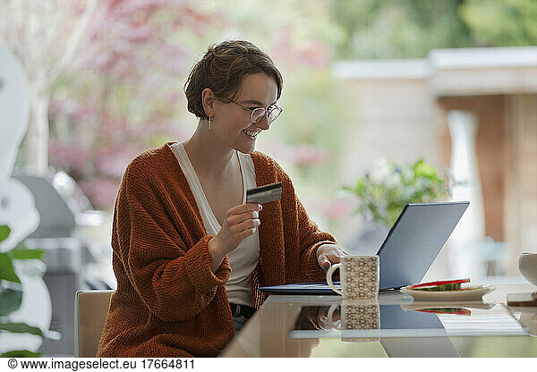 Young woman with credit card paying bills online at laptop in kitchen