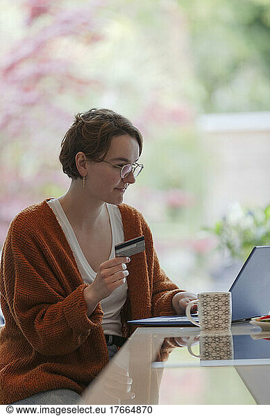 Young woman with credit card paying bills online at laptop in kitchen