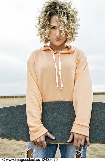Young woman with curly blond hair wearing pink hoodie and ripped jeans standing on sandy beach  holding skateboard  looking at camera.