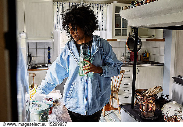 Young woman with curly hair holding drinking glasses while standing in kitchen at home
