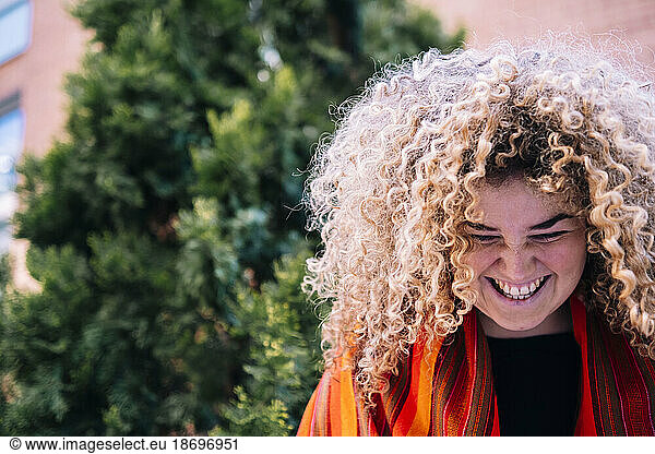 Young woman with curly hair laughing in front of tree