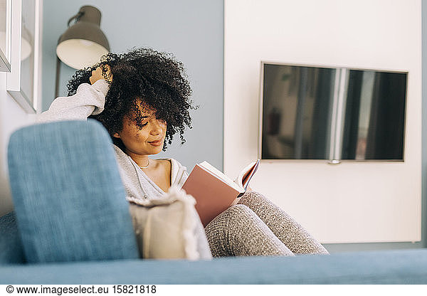 Young woman with curly hair reading a book on sofa at home