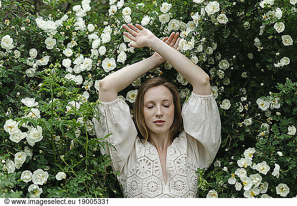 Young woman with eyes closed and arms raised by white flower bush