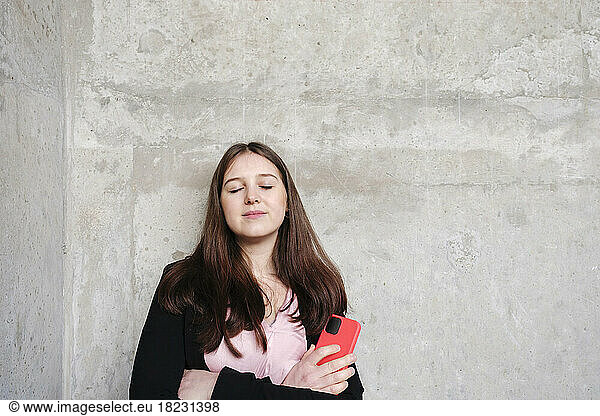 Young woman with eyes closed holding mobile phone leaning on wall