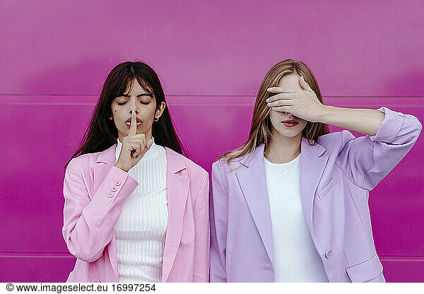 Young woman with finger on lips standing by sister covering eyes against pink wall