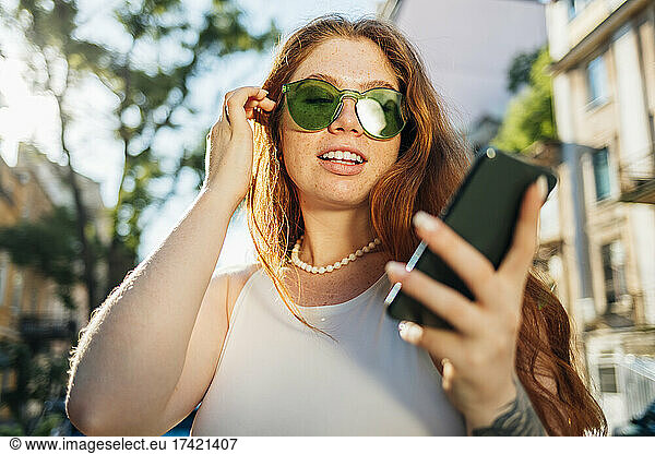 Young woman with hand in hair wearing green sunglasses using smart phone