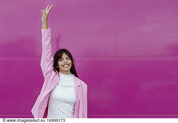 Young woman with hand raised doing peace sign while standing against pink wall