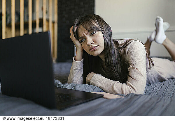 Young woman with head in hand using laptop on bed at home