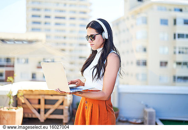 Young woman with headphones and laptop working on urban rooftop