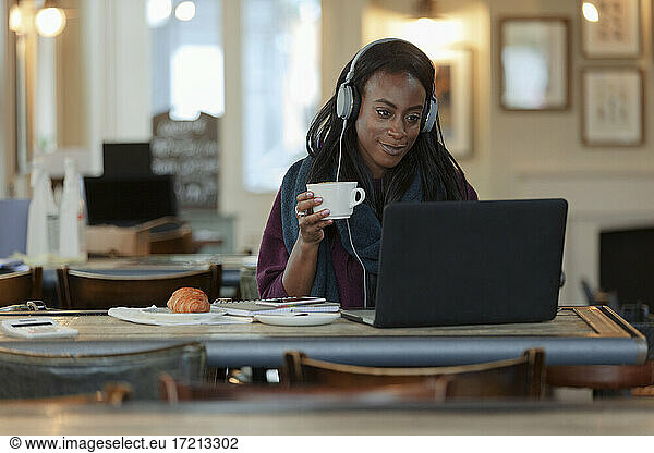 Young woman with headphones drinking coffee at laptop in cafe