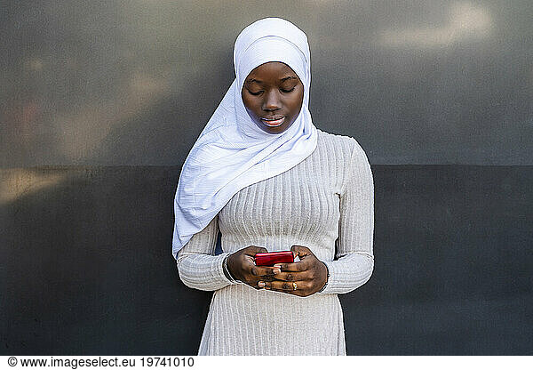Young woman with hijab using smart phone in front of wall