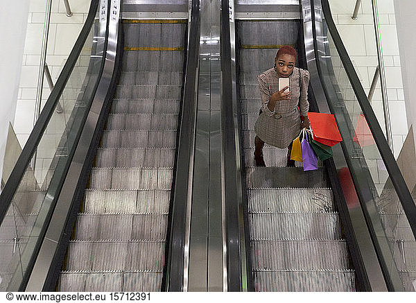 Young woman with holding colorful shopping bags and using her phone while standing on escalator