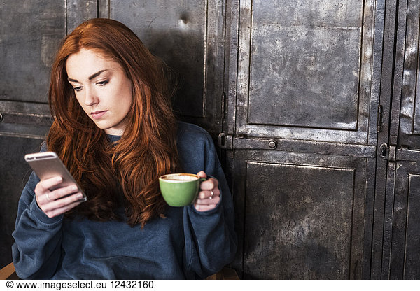 Young woman with long red hair sitting at table  holding cup of coffee  checking her mobile phone.