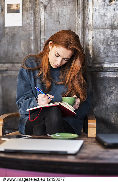 Young woman with long red hair sitting at table  holding notebook and cup of coffee.