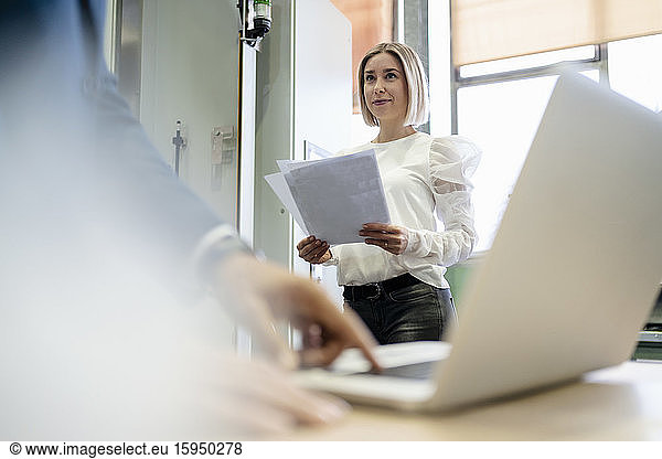 Young woman with papers looking at businessman using laptop in a factory