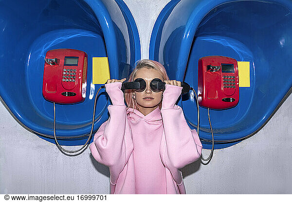 Young woman with pink hair wearing pink hooded shirt standing at telephone booths  holding receivers in front of eyes