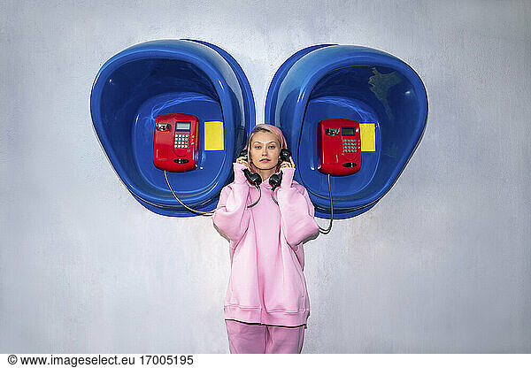 Young woman with pink hair wearing pink hooded shirt standing in front of telephone booths and holding receivers